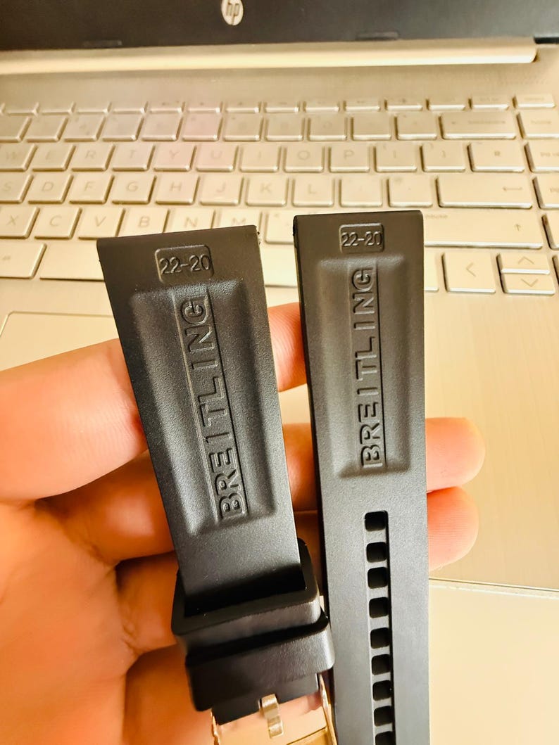 Puede incluir: Dos correas de reloj negras con el nombre de la marca "BREITLING" en relieve y la talla "22-20". Las correas se muestran frente a un teclado de ordenador port&aacute;til.