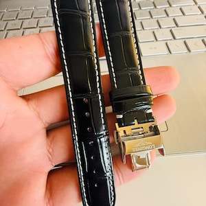 May include: Black watch strap with a crocodile pattern and white stitching. The strap features a silver clasp with the "LONGINES" brand name. The watch strap is made of a glossy material.