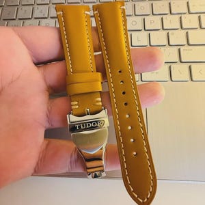May include: Mustard yellow leather watch strap with white stitching. The strap features a silver-colored buckle with the brand name "TUDOR" engraved on it. The watch strap is designed for a classic and elegant look.