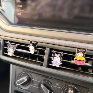 Könnte beinhalten: Vier Cartoon-Charakter-Autolüftungsclips. Die Clips sind schwarz, weiß, rosa und gelb. Die Clips sind an den Lüftungsschlitzen des Armaturenbretts eines Autos befestigt. Das Armaturenbrett ist schwarz und grau.