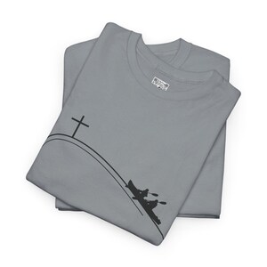 May include: Two gray t-shirts are folded and stacked. Each shirt features a black graphic of a cross on a hill and a kayak with two people paddling. The shirts are made of a soft material.