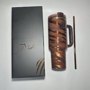 Puede incluir: Un vaso marr&oacute;n y dorado con asa y pajita a juego, junto a una caja negra con el texto "TYLA" y un dise&ntilde;o de hoja de palmera. El vaso tiene un estampado de rayas de tigre.