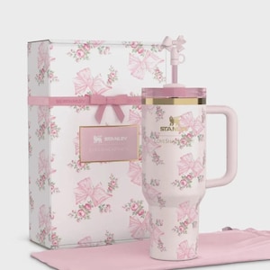 Puede incluir: Un vaso Stanley Loveshack Fancy rosa con asa, pajita y tapa, presentado con una caja de regalo a juego y una bolsa rosa. El vaso y la caja est&aacute;n adornados con un estampado floral y de lazo. La caja tiene una cinta rosa y una etiqueta.