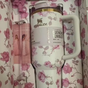 Puede incluir: Un vaso Stanley Quencher blanco con un estampado floral de rosas, un asa blanca y una tapa rosa. El vaso est&aacute; en una caja con un dise&ntilde;o floral a juego e incluye una pajita rosa. El texto en el vaso dice "QUENCHER TUMBLER 1.18L".