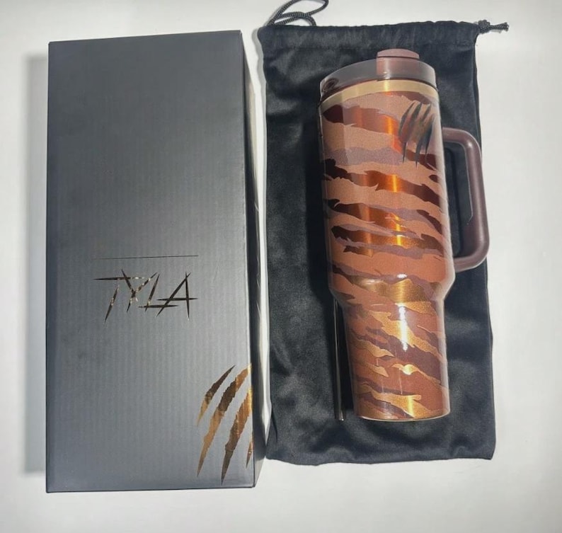 Puede incluir: Un termo de viaje marr&oacute;n y cobrizo con asa y tapa, con un dise&ntilde;o de rayas de tigre. El termo est&aacute; junto a una caja negra con el texto "TMA" y un dise&ntilde;o de garras. Tambi&eacute;n se incluye una bolsa negra con cord&oacute;n.