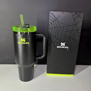 May include: Black Stanley travel mug with a green lid and straw, and a matching box. The mug has a textured pattern and the Stanley logo in yellow. The box features a spiderweb design.