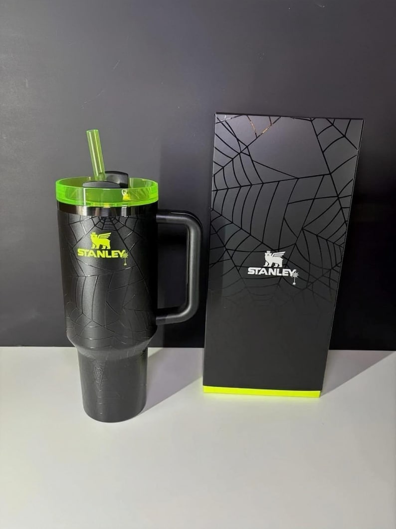 May include: Black Stanley travel mug with a green lid and straw, and a matching box. The mug has a textured pattern and the Stanley logo in yellow. The box features a spiderweb design.