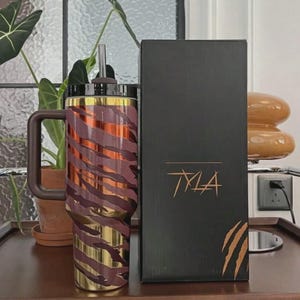 Puede incluir: Un vaso de acero inoxidable con asa, pajita y tapa. El vaso tiene una base dorada y est&aacute; decorado con un estampado de rayas de tigre marr&oacute;n y dorado. Una caja negra con la palabra "TYLA" en letras doradas est&aacute; al lado del vaso.