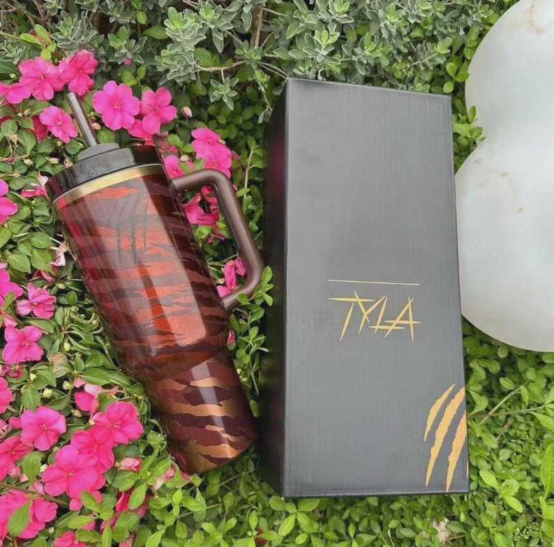 Puede incluir: Un vaso de color marr&oacute;n y marr&oacute;n con asa y pajita, con un estampado de rayas de tigre. El vaso est&aacute; junto a una caja negra con la palabra "TYLA" en letras doradas y un dise&ntilde;o de garra. Los art&iacute;culos est&aacute;n colocados entre flores rosas y vegetaci&oacute;n.