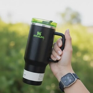May include: A black Stanley insulated mug with a green lid and handle. The mug has the Stanley logo in green. The mug is held by a person. A smartwatch is on the person's wrist.