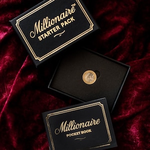 Puede incluir: Un juego de cajas negras con letras doradas que dice "Millionaire STARTER PACK" y un "Millionaire POCKET BOOK". Dentro de la caja hay una moneda de oro y plata. Los artículos están sobre una superficie de terciopelo burdeos.