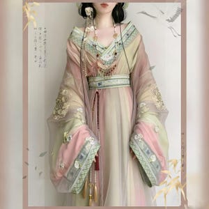 Puede incluir: Un vestido tradicional fluido y multicolor con mangas largas y transparentes. El vestido presenta bordados intrincados, un cinturón decorativo y un collar largo de cuentas. La paleta de colores incluye rosas, verdes y cremas suaves.