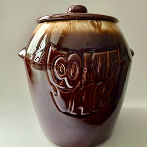 Pode incluir: Um pote de biscoitos vintage de cerâmica marrom com tampa. O pote tem um acabamento brilhante e apresenta um design em relevo com a palavra "Cookies". A tampa tem um pequeno botão para facilitar a remoção. O pote tem duas pequenas alças nas laterais.