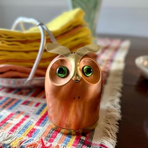 May include: A copper-colored owl figurine with brass accents and green glass eyes. The owl has a hinged base and a decorative design. It sits on a colorful fringed table runner. A basket of yellow napkins is in the background.