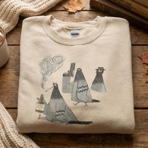 Puede incluir: Sudadera beige de cuello redondo con una ilustración humorística de tres palomas. Una paloma fuma un cigarrillo y sostiene una botella, otra sostiene una pistola y la tercera lleva un sombrero. El diseño es en tonos grises y negros.