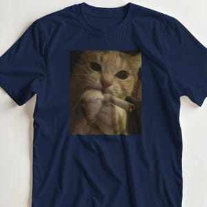 Puede incluir: Camiseta azul marino con una imagen de un gato sosteniendo un cigarrillo encendido. El gato tiene pelaje de color naranja claro y mira directamente al espectador. El cigarrillo est&aacute; encendido y tiene un brillo rojo en la punta.