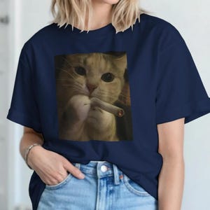 Puede incluir: Camiseta azul marino con una imagen humor&iacute;stica de un gato sosteniendo un cigarrillo encendido. El gato tiene pelaje marr&oacute;n y blanco y mira directamente al espectador. La imagen est&aacute; centrada en la camiseta.