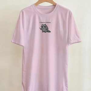 Puede incluir: Camiseta rosa claro con un dibujo de un mapache sosteniendo un cuchillo y el texto "I require snacks". El mapache es en blanco y negro. La camiseta tiene mangas cortas y cuello redondo.