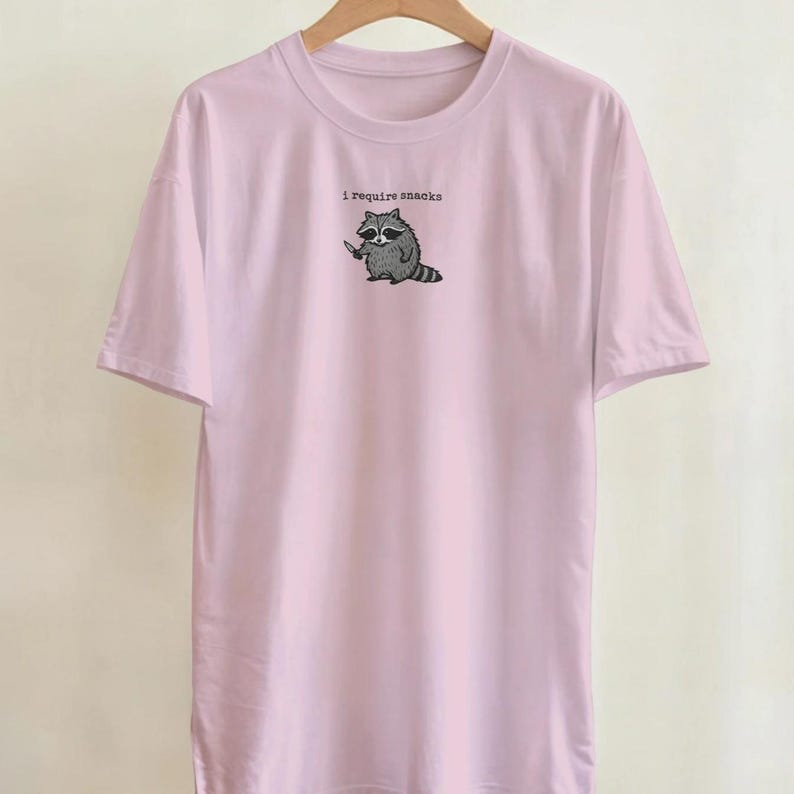 Puede incluir: Camiseta rosa claro con un dibujo de un mapache sosteniendo un cuchillo y el texto "I require snacks". El mapache es en blanco y negro. La camiseta tiene mangas cortas y cuello redondo.