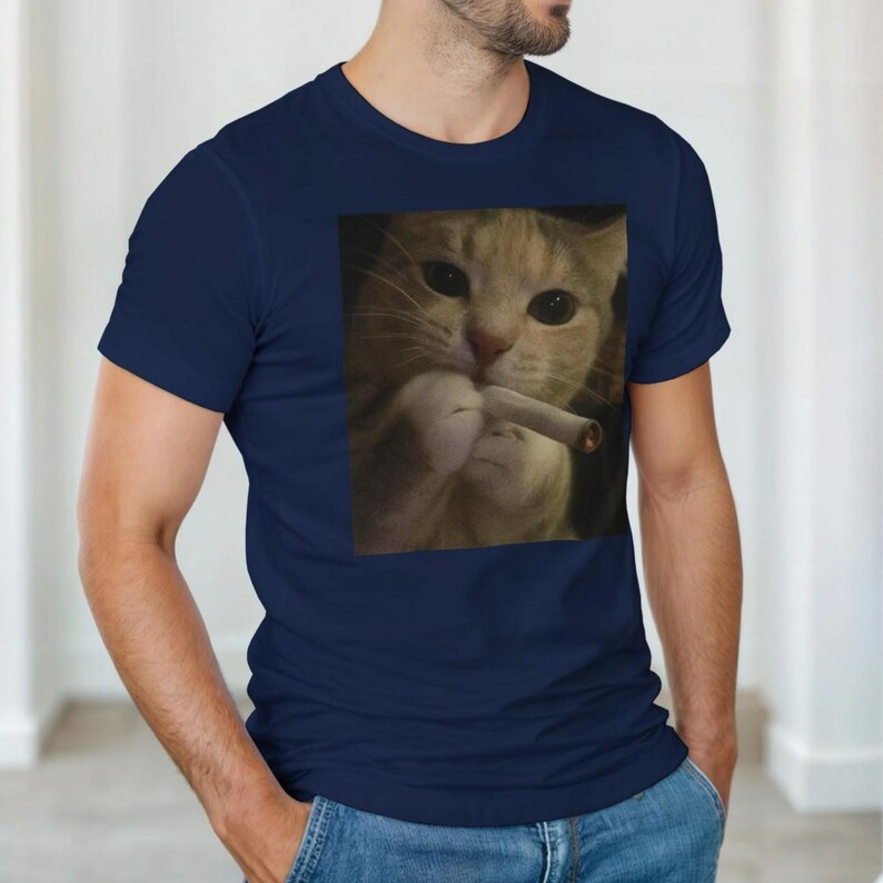 Puede incluir: Camiseta azul marino con una imagen de un gato sosteniendo un cigarrillo. El gato tiene pelaje de color naranja claro y blanco. La camiseta es de color liso y tiene cuello redondo. El gato mira directamente al espectador.