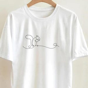 May include: White t-shirt with a minimalist black line drawing of a squirrel. The design shows a squirrel holding a nut, with a curved line extending from its body. The t-shirt is displayed on a wooden hanger.