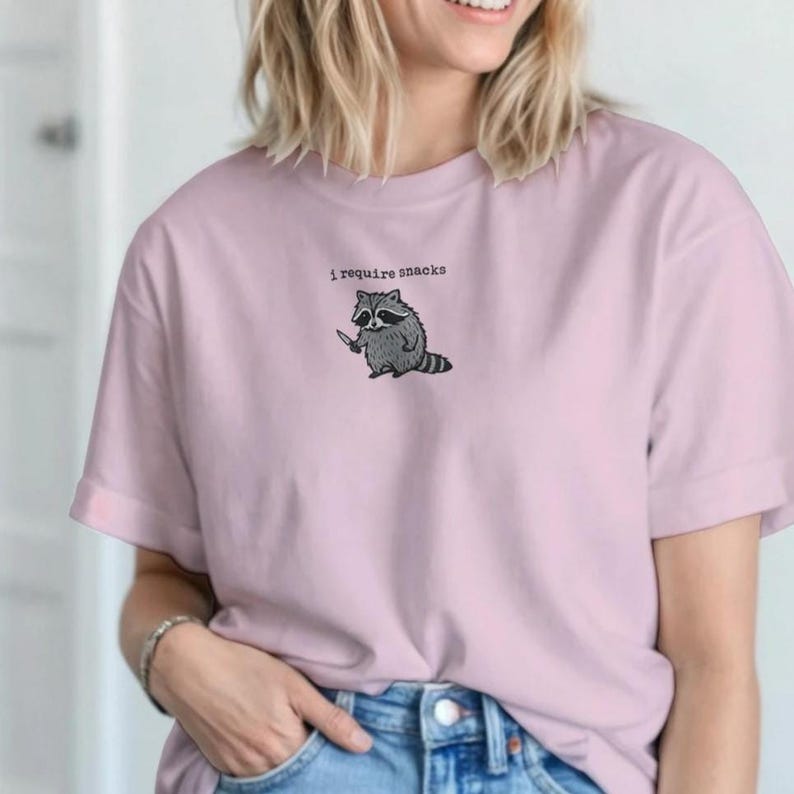 Puede incluir: Camiseta rosa claro con la frase "I require snacks" sobre un mapache de dibujos animados sosteniendo un cuchillo. El mapache es gris y blanco con una m&aacute;scara y cola negras. La camiseta tiene mangas cortas.