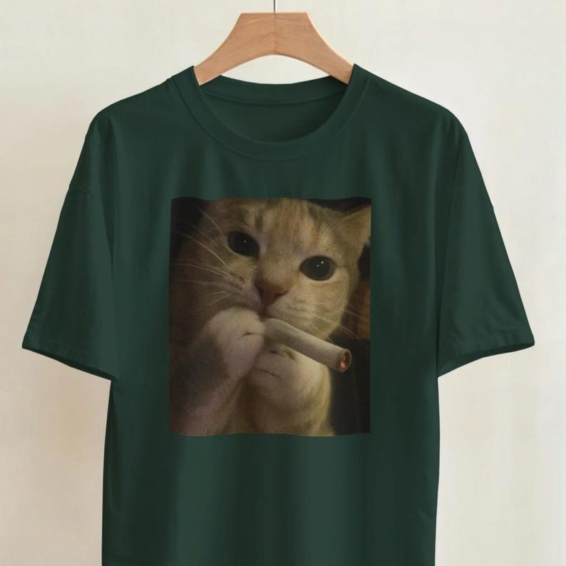 Puede incluir: Camiseta verde oscuro con una imagen de un gato sosteniendo un cigarrillo. El gato tiene pelaje naranja y blanco y mira directamente al espectador. La camiseta est&aacute; colgada en una percha de madera.
