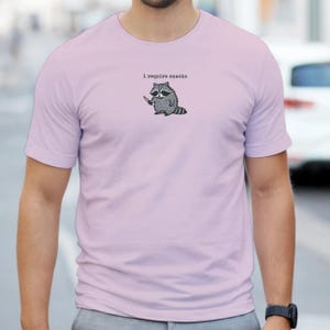 Puede incluir: Camiseta de color lila claro con un mapache de dibujos animados sosteniendo un cuchillo. El texto "i require snacks" est&aacute; encima del mapache. El mapache es gris y blanco con un contorno negro. La camiseta es de manga corta.