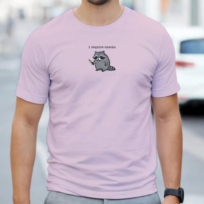 Puede incluir: Camiseta de color lila claro con un mapache de dibujos animados sosteniendo un cuchillo. El texto "i require snacks" est&aacute; encima del mapache. El mapache es gris y blanco con un contorno negro. La camiseta es de manga corta.