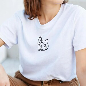 以下が含まれることがあります： 白のTシャツ。黒い線で描かれたミニマリストなキツネの絵が特徴です。キツネは座った姿勢で、尻尾が丸まっています。Tシャツは半袖で、クルーネックです。
