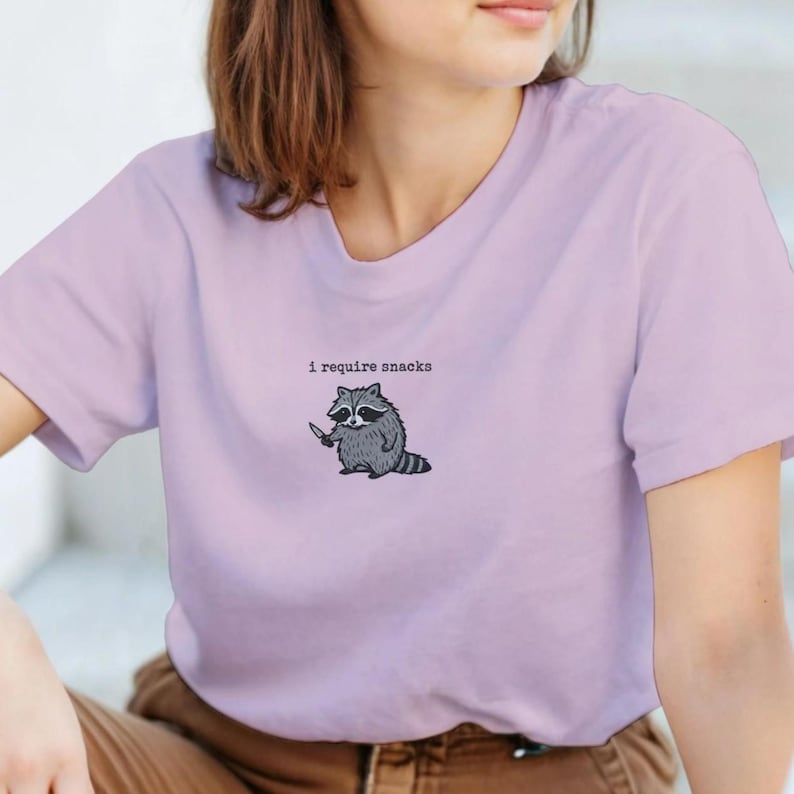 Puede incluir: Camiseta lila claro con un mapache de dibujos animados sosteniendo un cuchillo. El texto "i require snacks" est&aacute; encima del mapache. La camiseta tiene mangas cortas y cuello redondo.