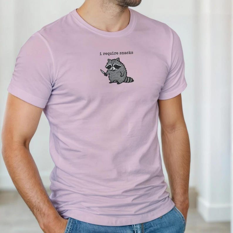Puede incluir: Camiseta de color lila claro con un mapache de dibujos animados sosteniendo un cuchillo. El mapache es en blanco y negro, y el texto "i require snacks" est&aacute; encima del mapache.