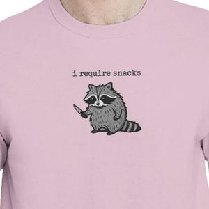 Puede incluir: Camiseta rosa claro con un dibujo de un mapache sosteniendo un cuchillo. El mapache es gris y negro, con una cola a rayas. El texto "i require snacks" est&aacute; impreso arriba.