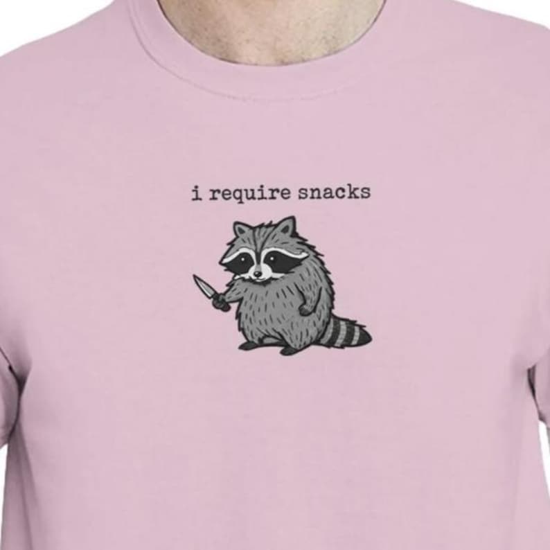 Puede incluir: Camiseta rosa claro con un dibujo de un mapache sosteniendo un cuchillo. El mapache es gris y negro, con una cola a rayas. El texto "i require snacks" est&aacute; impreso arriba.