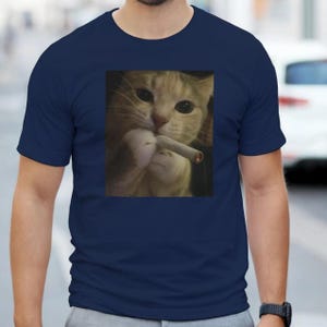 Puede incluir: Camiseta azul marino con una imagen de un gato sosteniendo un cigarrillo. El gato tiene pelaje naranja y blanco y mira al espectador. La camiseta est&aacute; hecha de un material suave.