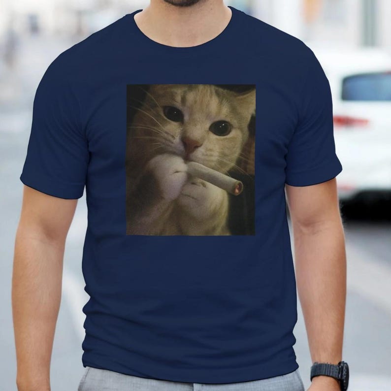Puede incluir: Camiseta azul marino con una imagen de un gato sosteniendo un cigarrillo. El gato tiene pelaje naranja y blanco y mira al espectador. La camiseta est&aacute; hecha de un material suave.