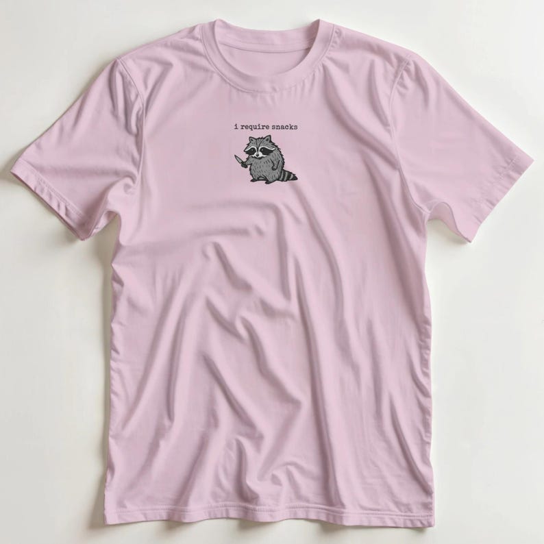 Puede incluir: Camiseta rosa claro con un dibujo de un mapache sosteniendo un cuchillo y el texto "I require snacks" encima. El mapache es en blanco y negro, y la camiseta es de color liso.