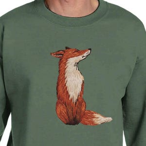Könnte beinhalten: Grünes Sweatshirt mit einem gestickten Fuchs. Der Fuchs ist in Braun-, Weiß- und Orangetönen dargestellt, mit einem detaillierten Design. Das Sweatshirt hat eine einfarbige grüne Farbe, und der Fuchs ist mittig auf der Vorderseite platziert.