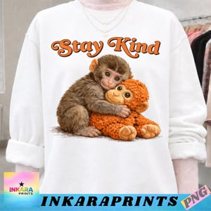 Puede incluir: Sudadera blanca con un gráfico de dos monos abrazándose, uno marrón y otro naranja. El texto "Stay Kind" está escrito sobre los monos en letras naranjas. La sudadera tiene mangas largas.