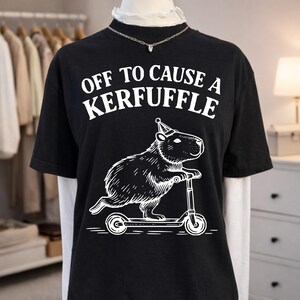 May include: Black t-shirt with the text "OFF TO CAUSE A KERFUFFLE" in white. Below the text is a white illustration of a capybara riding a scooter and wearing a party hat. The shirt is displayed on a mannequin.