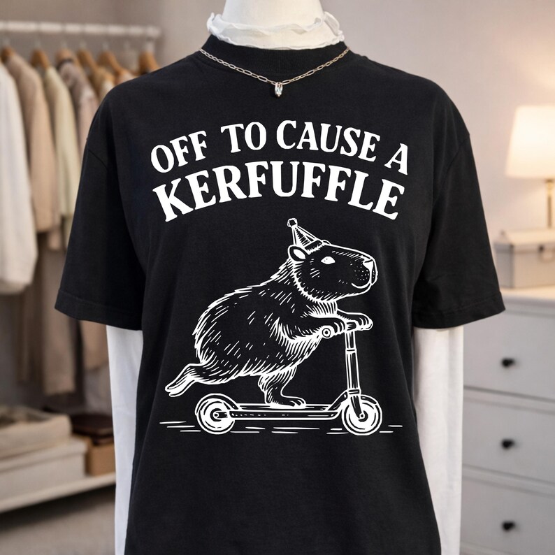 May include: Black t-shirt with the text "OFF TO CAUSE A KERFUFFLE" in white. Below the text is a white illustration of a capybara riding a scooter and wearing a party hat. The shirt is displayed on a mannequin.