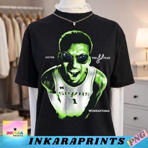 Pode incluir: Camiseta preta com um gráfico verde de um jogador de basquete com olhos alienígenas. O design inclui o texto "VICTOR", "THE 4 FILES" e "WEMBANYAMA". O jogador está vestindo uma camisa dos Spurs com o número 1.
