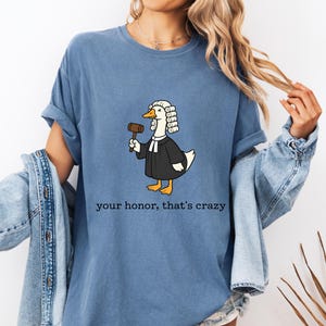 May include: Blue t-shirt featuring a cartoon duck dressed as a judge, holding a gavel. The duck wears a wig and a black robe. The text "your honor, that's crazy" is printed below the duck.