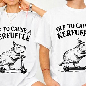 May include: White t-shirts with a black graphic of a capybara riding a scooter and wearing a party hat. The text "OFF TO CAUSE A KERFUFFLE" is printed above the capybara. The shirts are unisex.