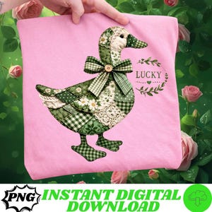 Peut inclure: T-shirt rose avec un motif d'oie en patchwork vert, blanc et crème. L'oie a un nœud et le mot "LUCKY" est écrit sur le t-shirt. Le t-shirt est tenu par une main.