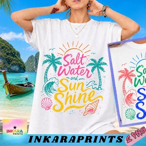 Puede incluir: Camiseta blanca con un diseño gráfico colorido con las palabras "Salt Water and Sun Shine". El diseño incluye palmeras, olas, un sol y conchas marinas. La camiseta es una prenda informal con temática de playa.