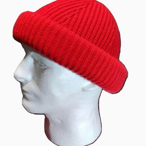 May include: A bright red beanie cap featuring a thick, ribbed knit texture and a wide, folded cuff. The hat is displayed on a white mannequin head, showcasing the vertical knit pattern that extends from the crown down to the brim.