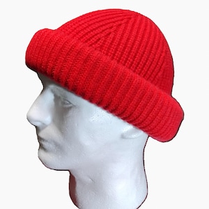 Puede incluir: Un gorro de punto acanalado de color rojo brillante. El gorro se muestra sobre una cabeza de maniquí blanca, destacando el borde doblado y el ajuste ceñido. La textura y el color del gorro destacan sobre el fondo blanco.
