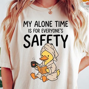 K&ouml;nnte beinhalten: Elfenbeinfarbenes T-Shirt mit dem Text "MY ALONE TIME IS FOR EVERYONE'S SAFETY". Ein Cartoon-Ente mit Handtuch und Bademantel liest ein Buch und h&auml;lt eine Kaffeetasse. Auf dem Shirt stehen auch die Worte "super soft" und "IVORY".
