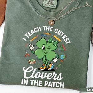 May include: Moss green t-shirt with the text "I TEACH THE CUTEST Clovers IN THE PATCH" and a cartoon clover graphic. The clover is wearing a bow and sneakers. The shirt has a crew neck and short sleeves.